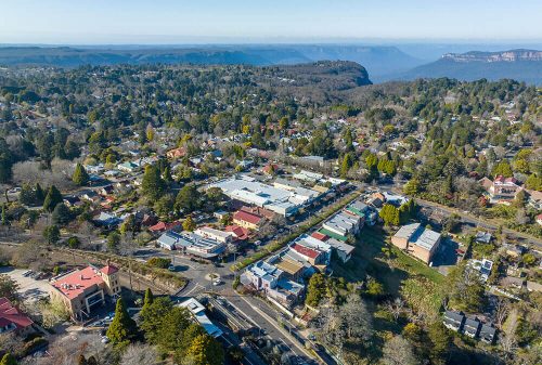 Aerial view of Leura Village with heritage buildings, tree-lined streets, and the vast Blue Mountains valley in the background.